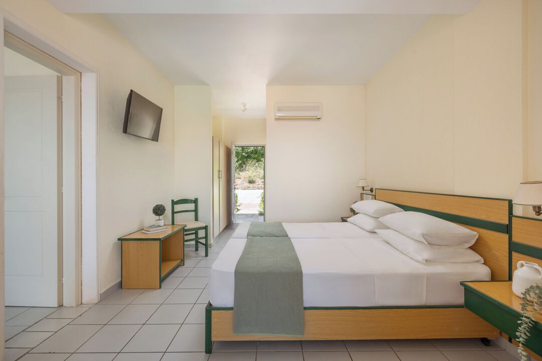 A bedroom with a bed, bedside table, chair, TV, and an air conditioning unit.