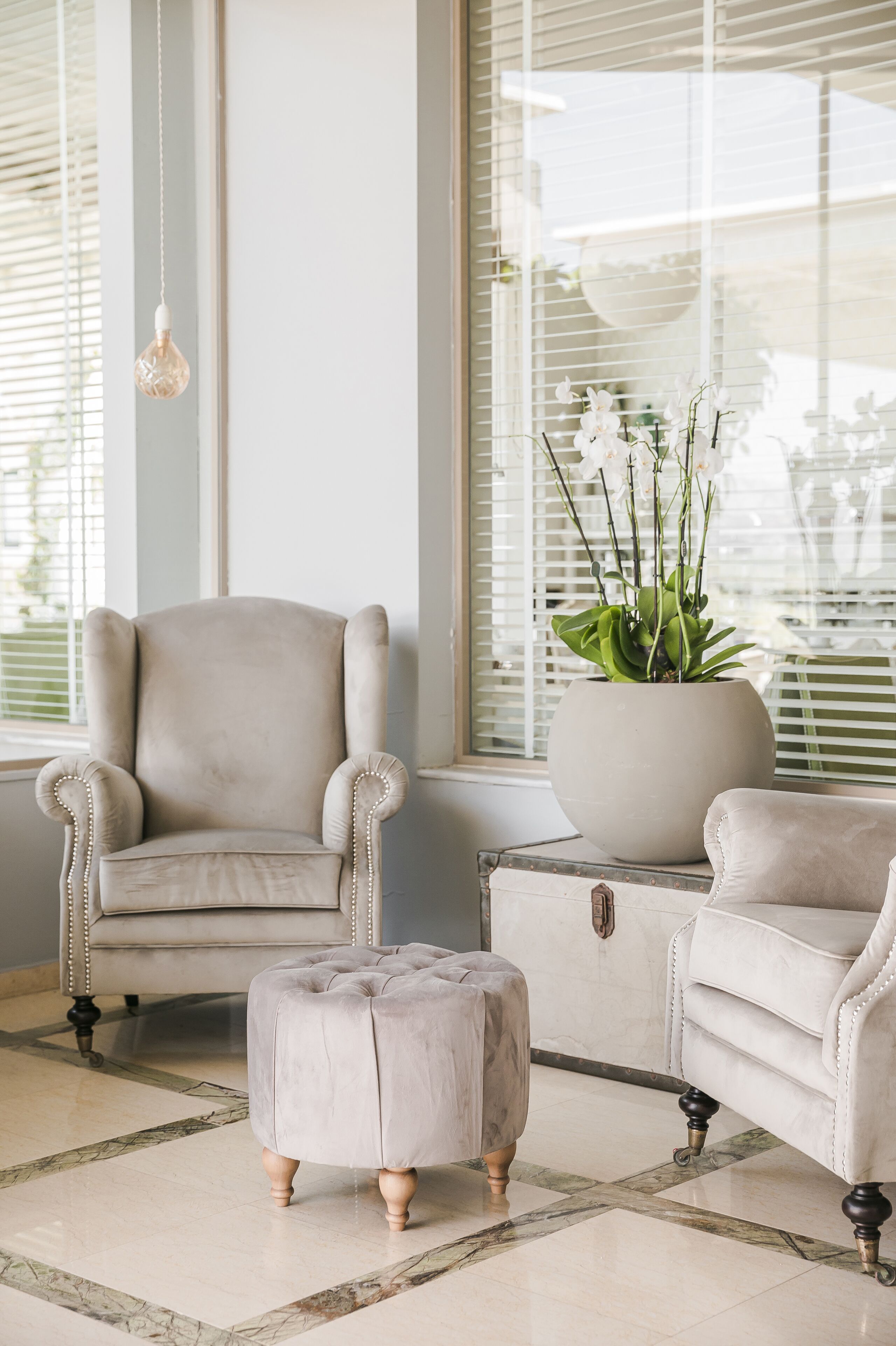 A room with a beige armchair, a matching ottoman, and a potted plant by a window with blinds.