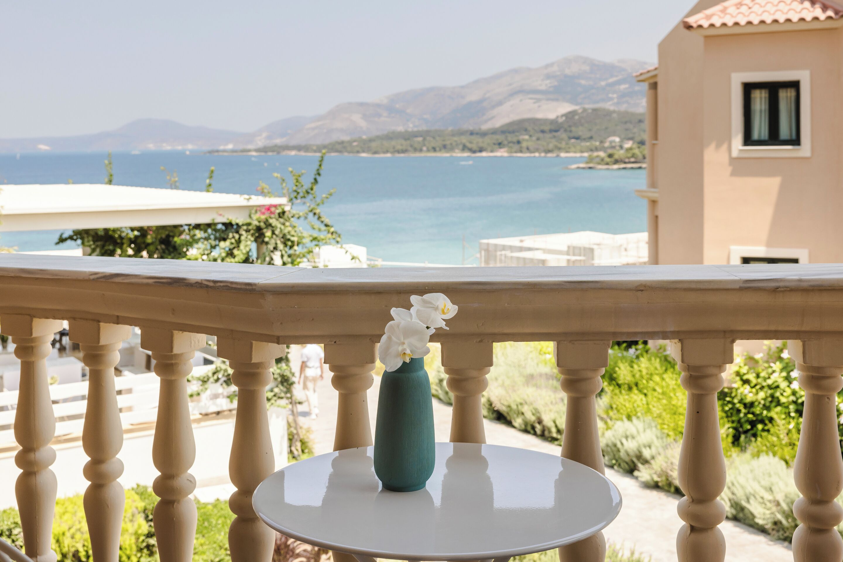 Ein Balkon mit einem weißen Tisch und einer Vase mit weißen Blumen, mit Blick auf die Küste und Berge in der Ferne.
