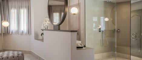 A modern bathroom with a glass shower enclosure, a round mirror, and a tufted ottoman.