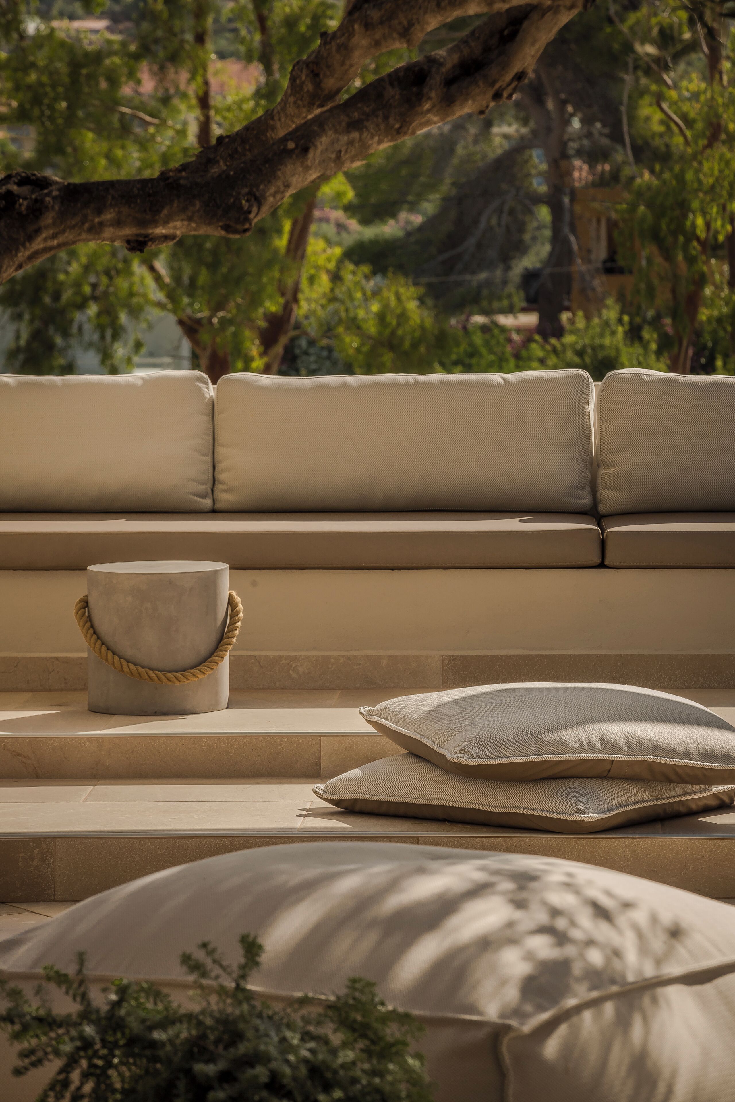 Un canapé d’extérieur de couleur beige avec des coussins, un pot de fleurs rond en béton doté d’une anse en corde, et un coussin posé sur le sol.