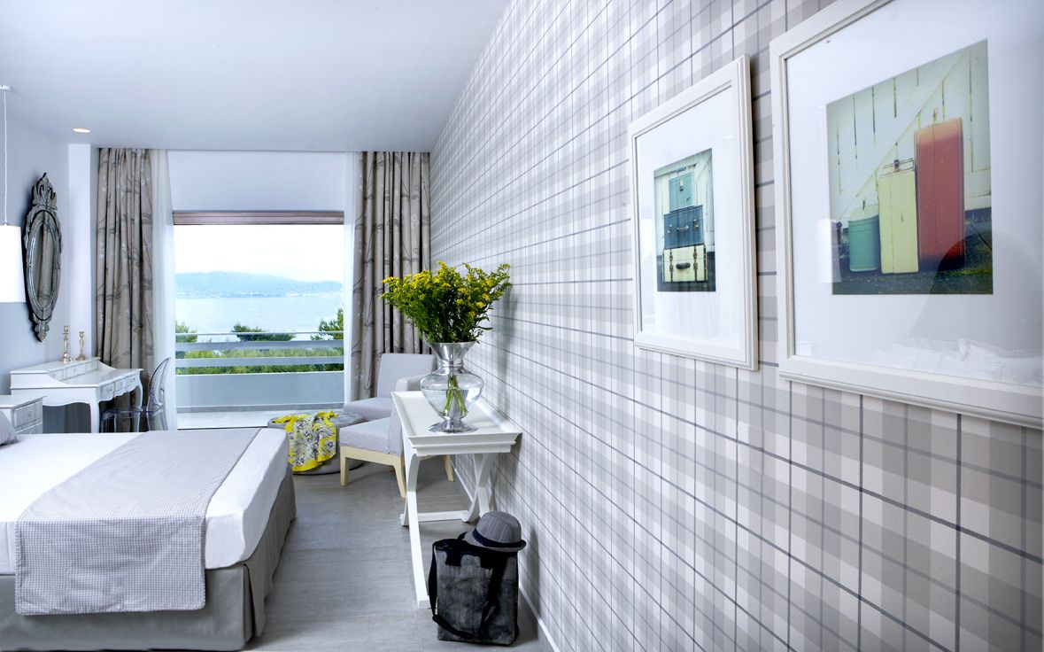A hotel room with a bed, a desk, a chair, a small table with a vase of flowers, and a view of the outdoors.
