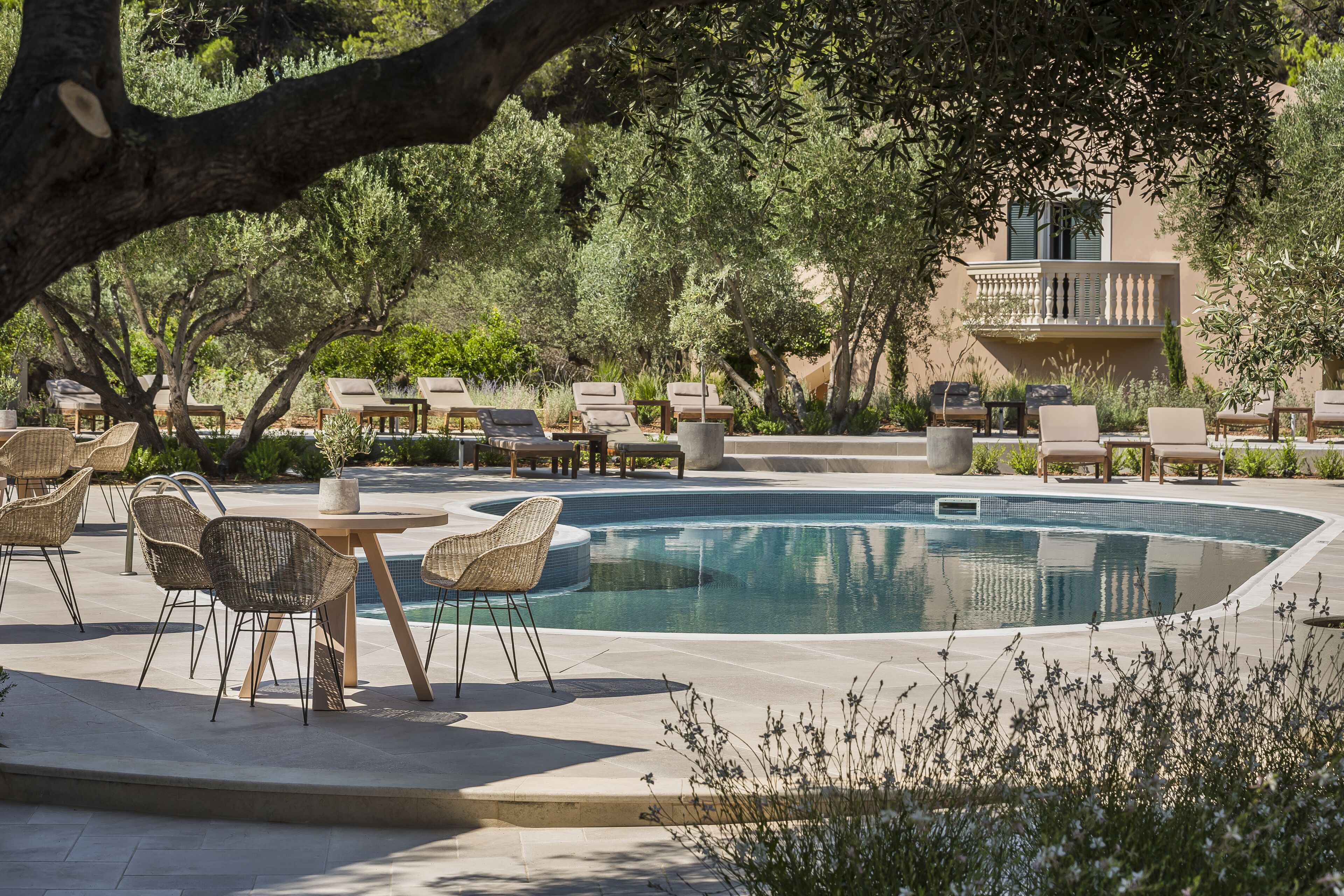A poolside area with lounge chairs and tables, surrounded by trees and a building with a balcony.