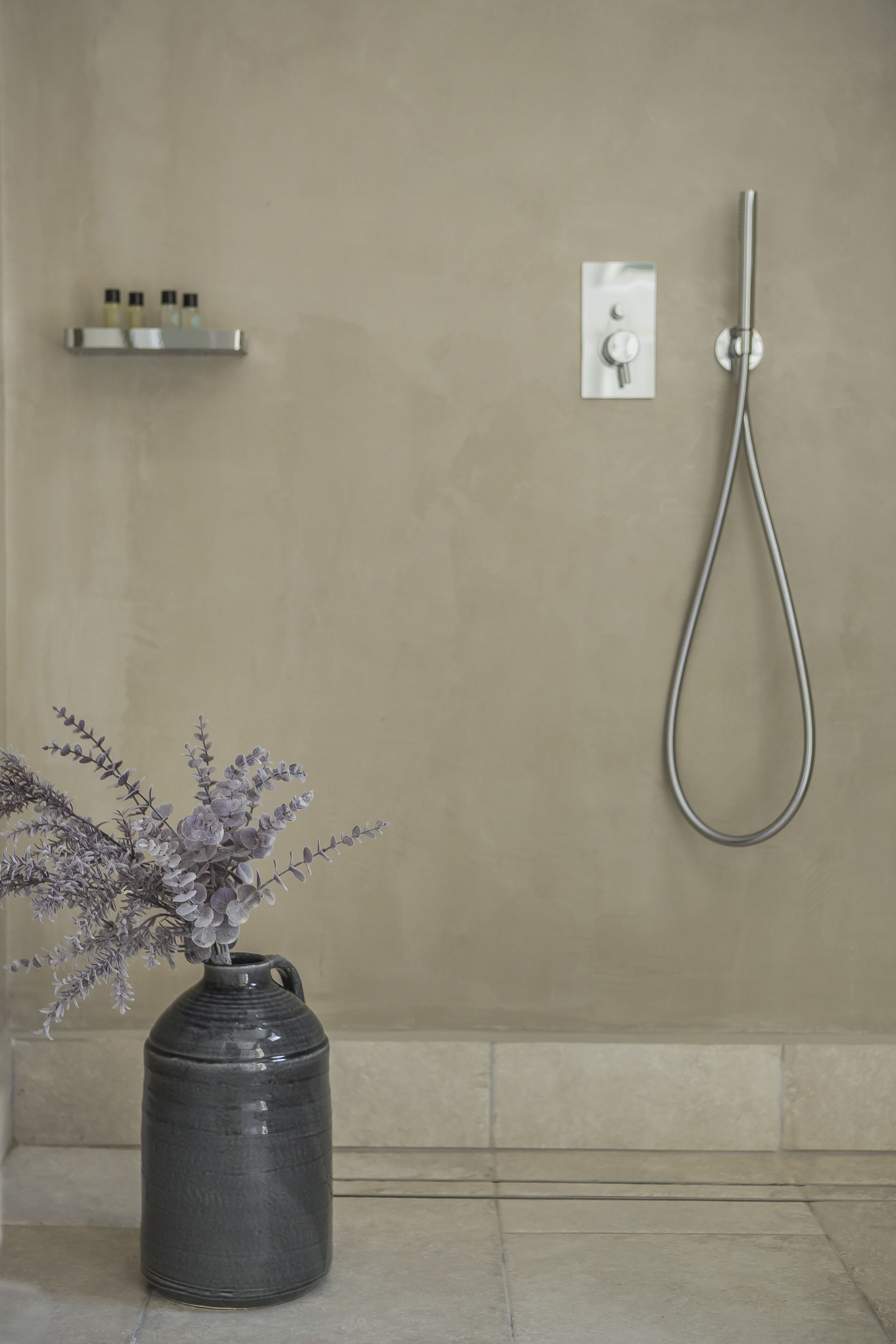 Une salle de bain avec un vase en céramique grise orné de fleurs, une douche et une petite étagère contenant des articles de toilette.
