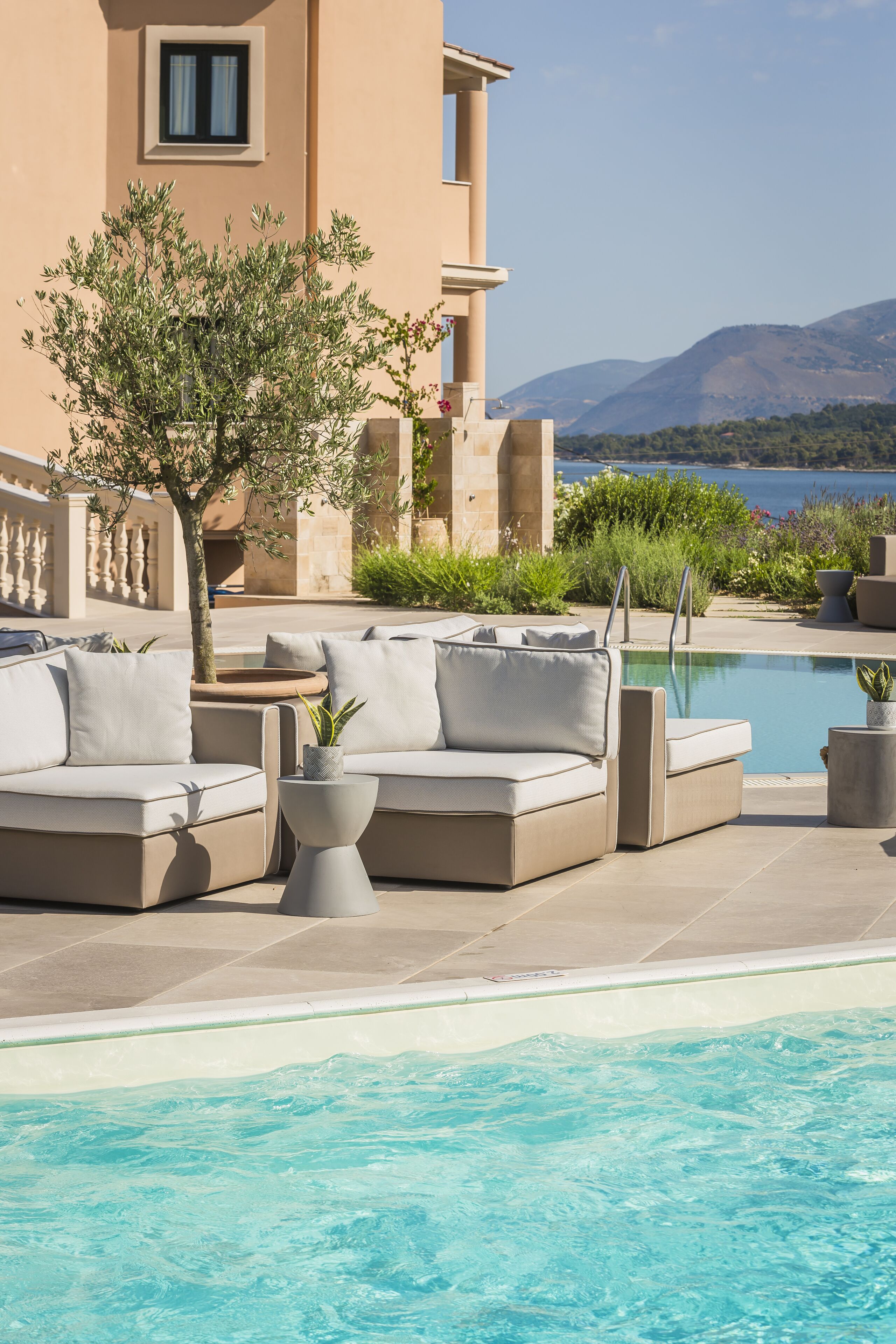 Zona junto a la piscina con un sofá color crema, un árbol pequeño y vistas al mar y a las montañas.
