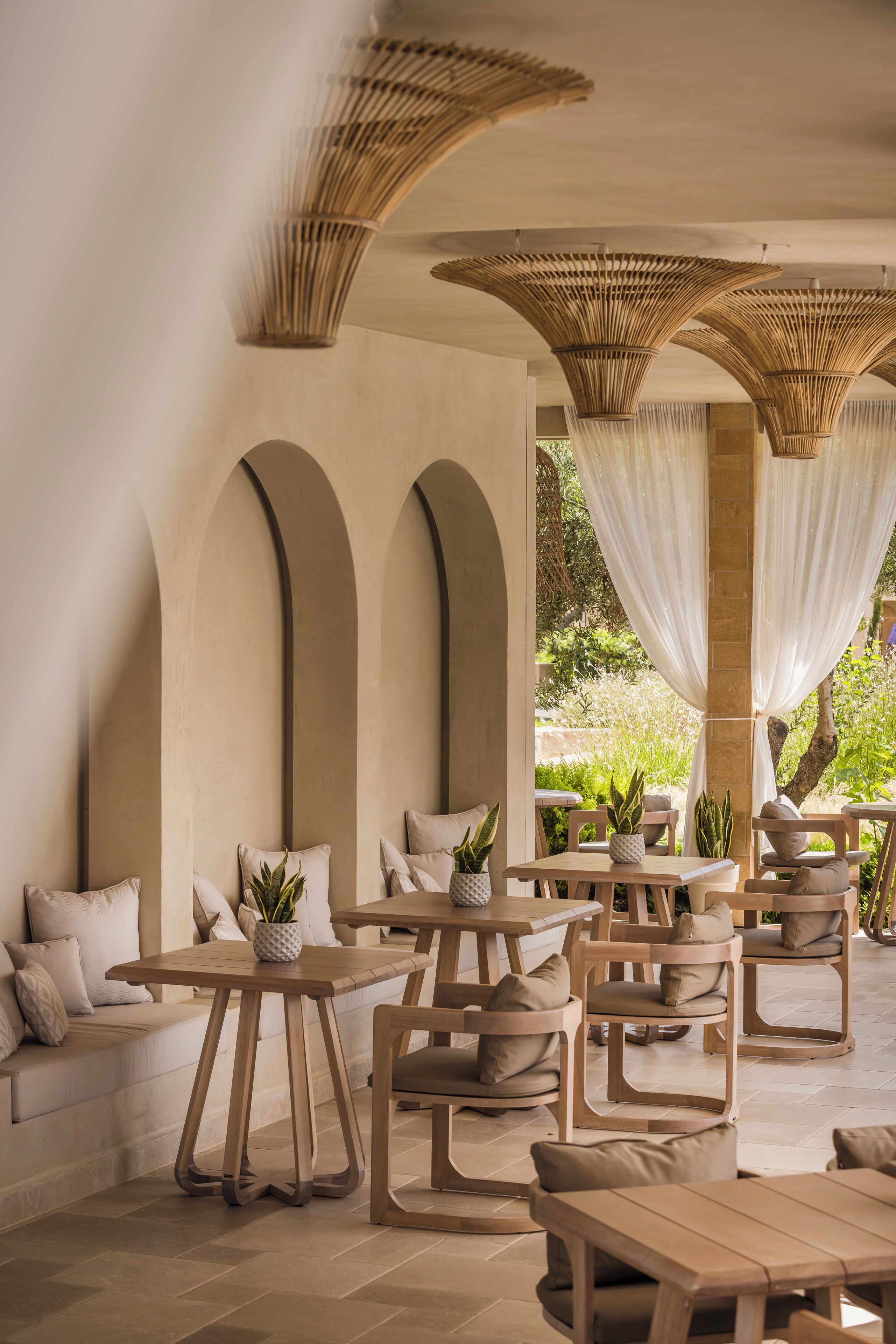 Un espace extérieur aménagé avec des tables et des chaises en bois, un banc en pierre et une pergola agrémentée de voilages blancs.