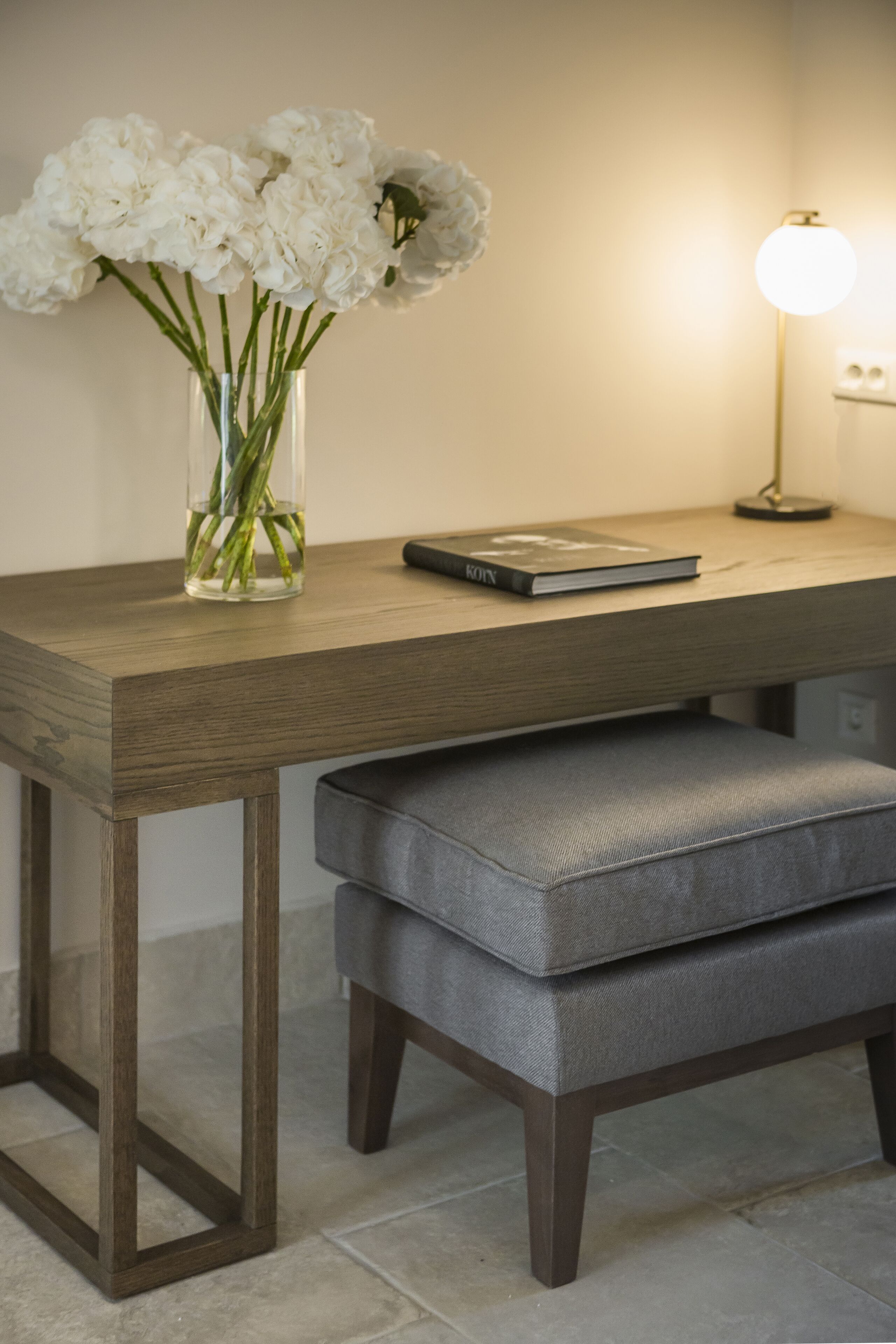 Un bureau en bois orné d’un vase de fleurs blanches, d’un livre et d’une lampe.