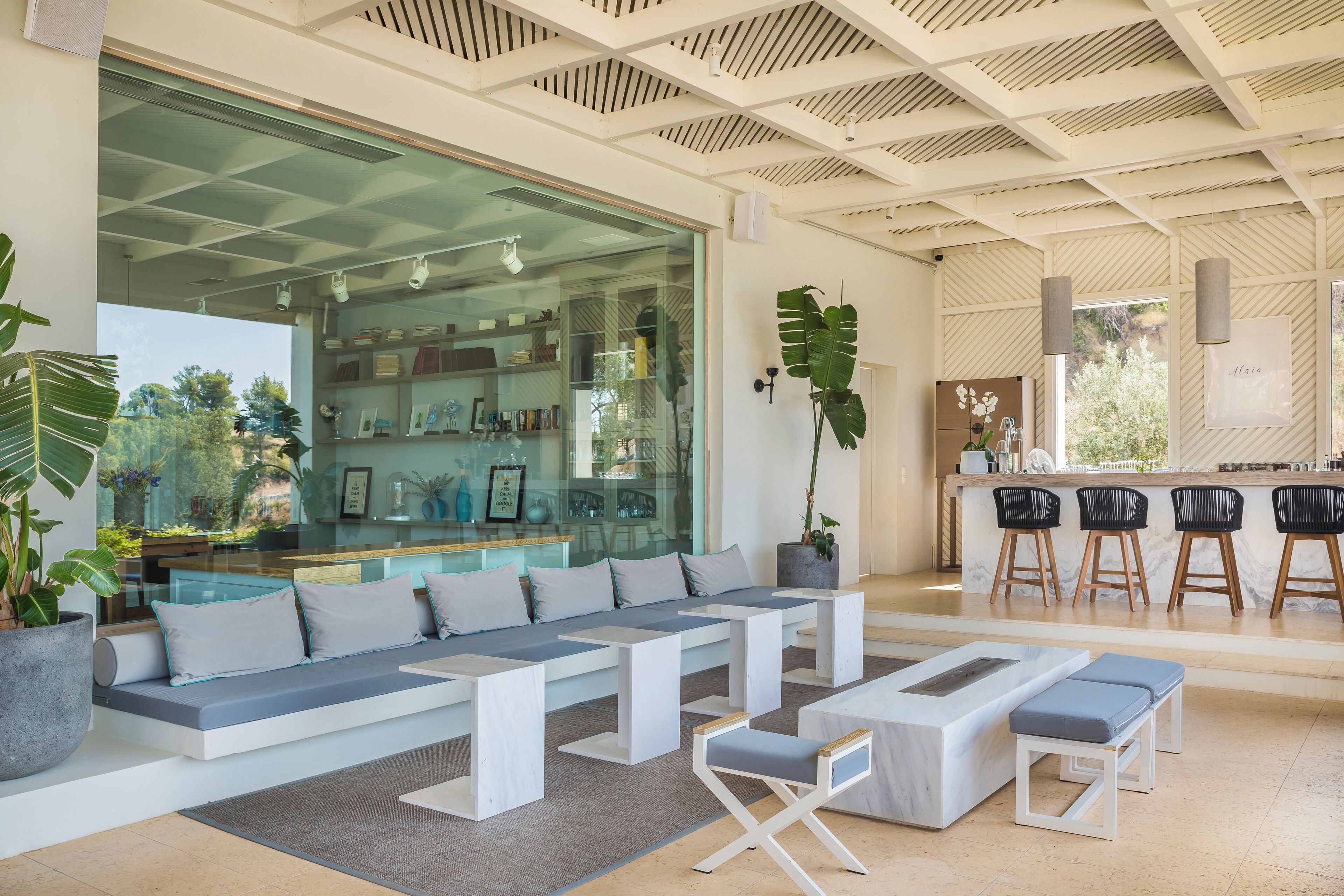 A modern living area with a large glass wall, a seating area with benches and cushions, a kitchen counter with bar stools, and a potted plant.