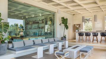 A modern living area with a large glass wall, a seating area with benches and cushions, a kitchen counter with bar stools, and a potted plant.
