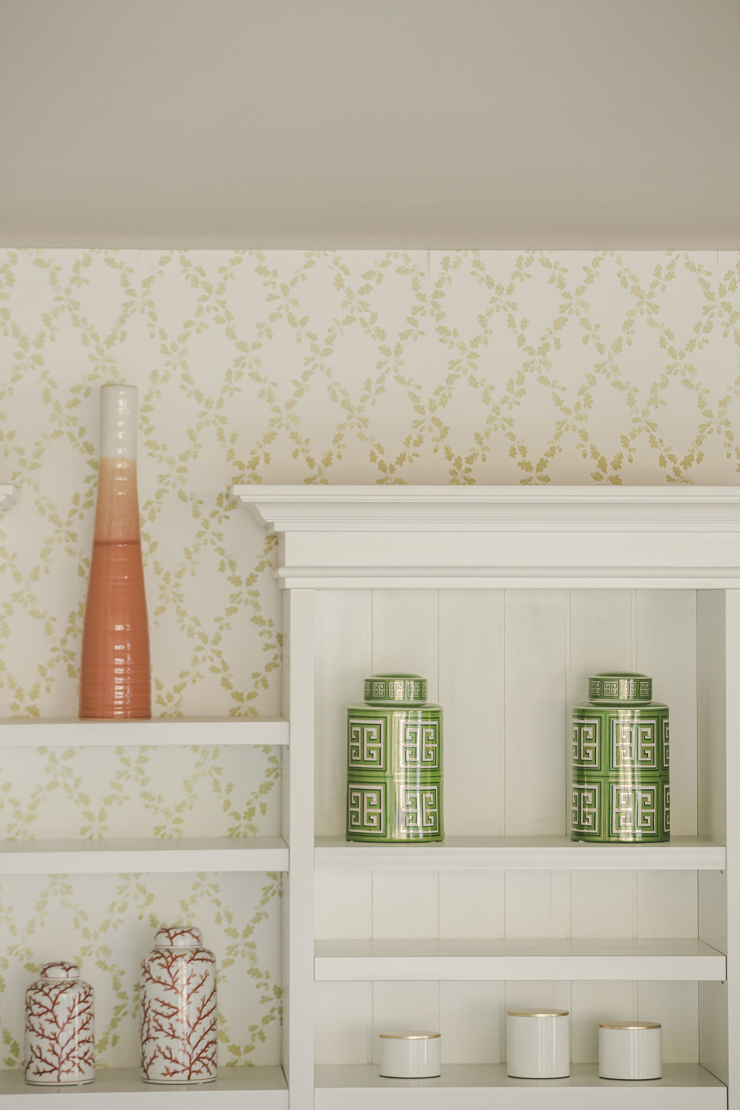 Une étagère blanche ornée de vases et de pots décoratifs, le tout placé devant un mur recouvert d’un papier peint à motifs.