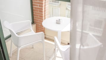Un balcón con sillas de mimbre blanco y una pequeña mesa redonda, una pared de ladrillo y una vista parcial de un edificio con barandillas blancas al fondo.