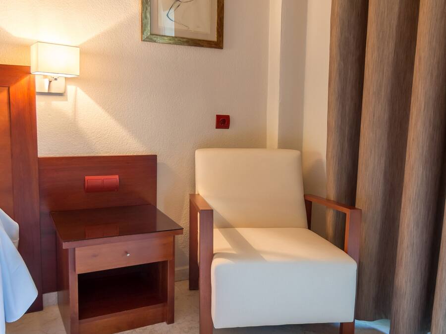 A hotel room with a wooden nightstand, a white chair, a framed artwork on the wall, and a lamp.