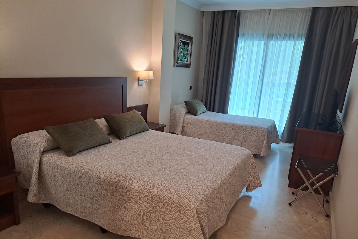 Una habitación de hotel con dos camas, una mesita de noche, una lámpara, una silla y una ventana con cortinas.