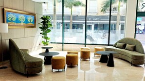 Un hall d’entrée moderne, aménagé avec un canapé, des fauteuils et des poufs disposés autour d’une table basse centrale. Une grande fenêtre offre une vue sur la rue et sur un immeuble appelé « EL YATE ».