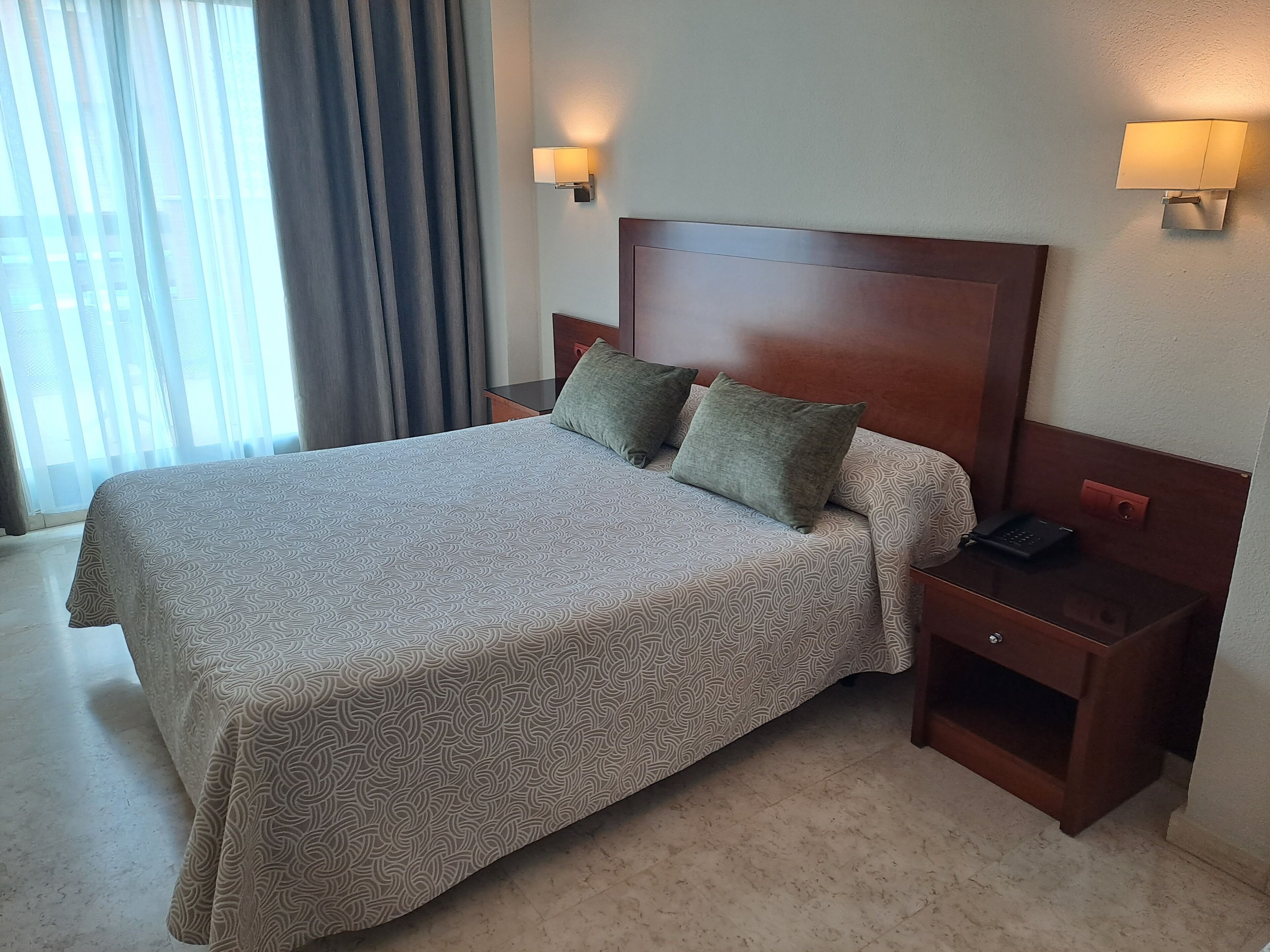 A hotel room with a large bed, two bedside tables, a nightstand, and a wall-mounted lamp.