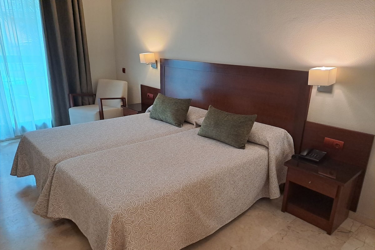 Una cama doble con una colcha estampada, dos almohadas, lámparas de noche, una mesita de noche con un teléfono y una silla en una habitación de hotel.