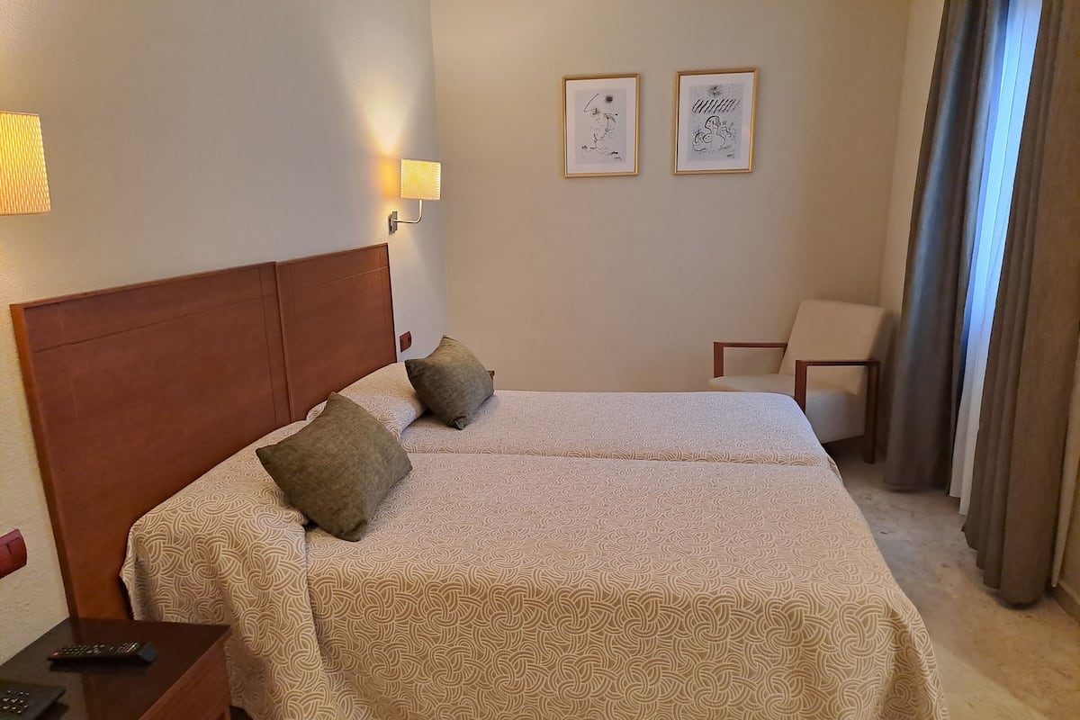 Habitación de hotel con cama doble, cabecero de madera, mesita de noche con control remoto, dos cuadros enmarcados en la pared y una silla con una mesa.