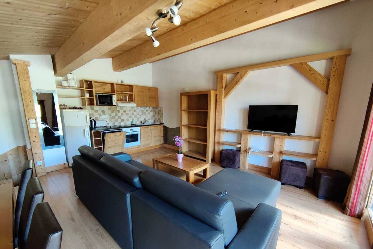 A modern living room with a sectional sofa, wooden beams, a television, and a kitchen area with wooden cabinets and appliances.