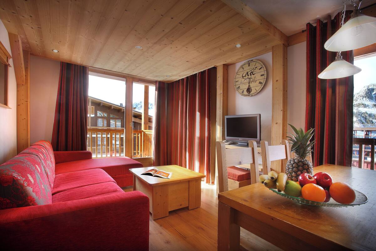 A cozy living room with a red sofa, wooden ceiling, a dining table with fruit, and a TV.
