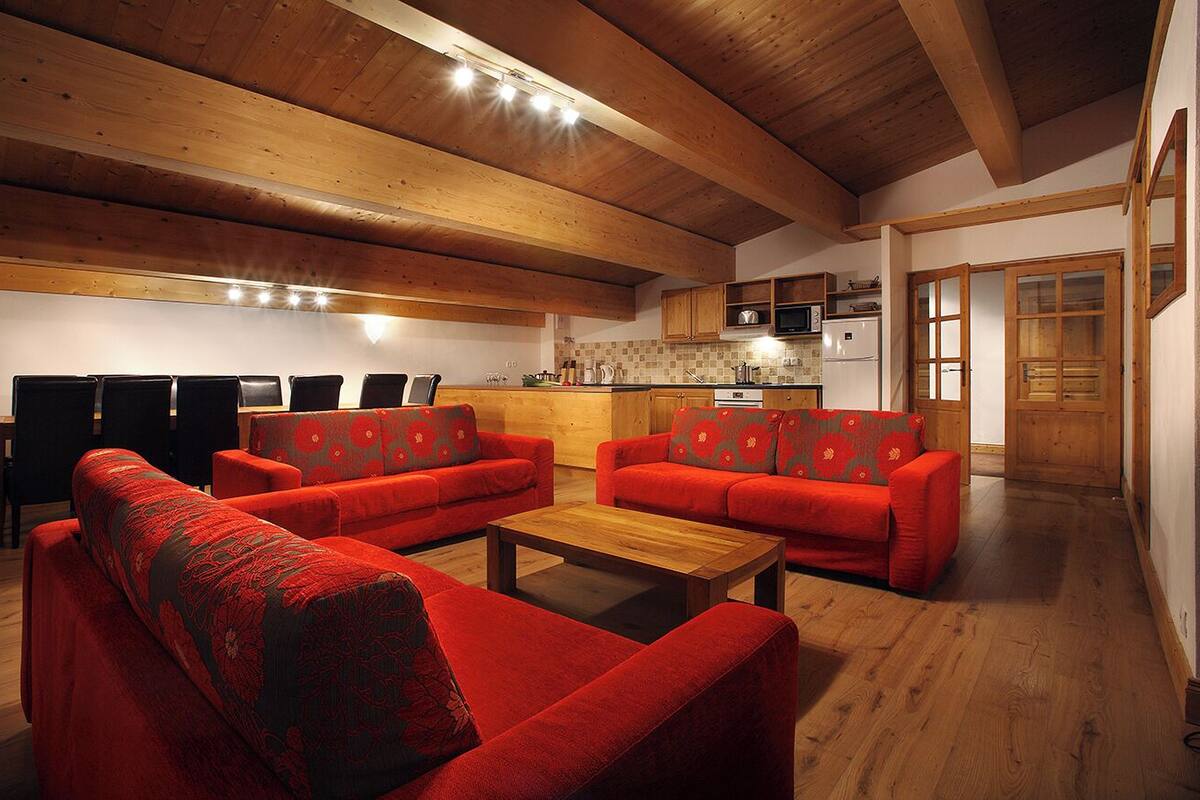 A spacious living area with red sofas and a wooden coffee table, a kitchen with modern appliances, and wooden beams on the ceiling.