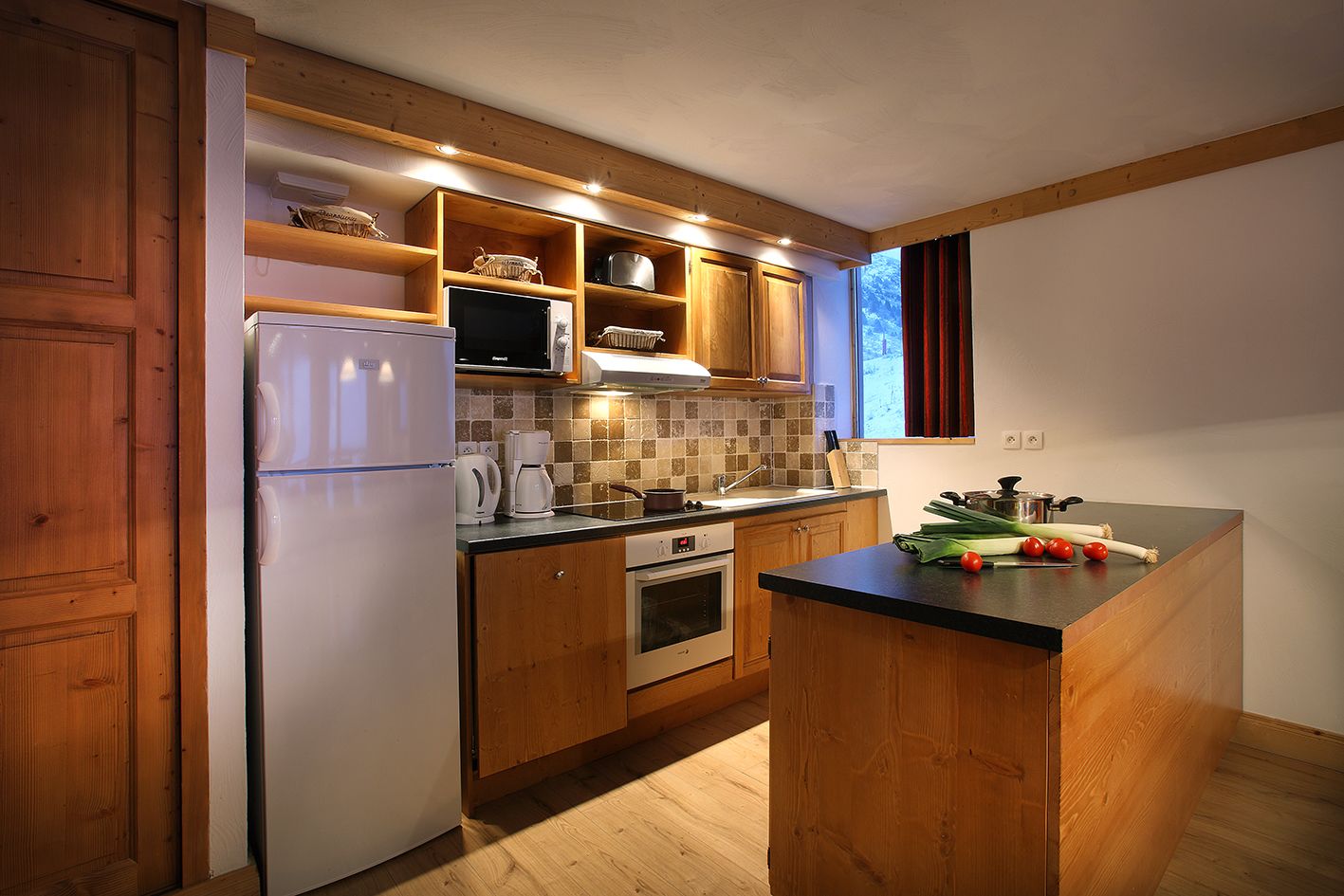Una cocina compacta con armarios de madera, horno empotrado, refrigerador y una pequeña isla con verduras frescas.