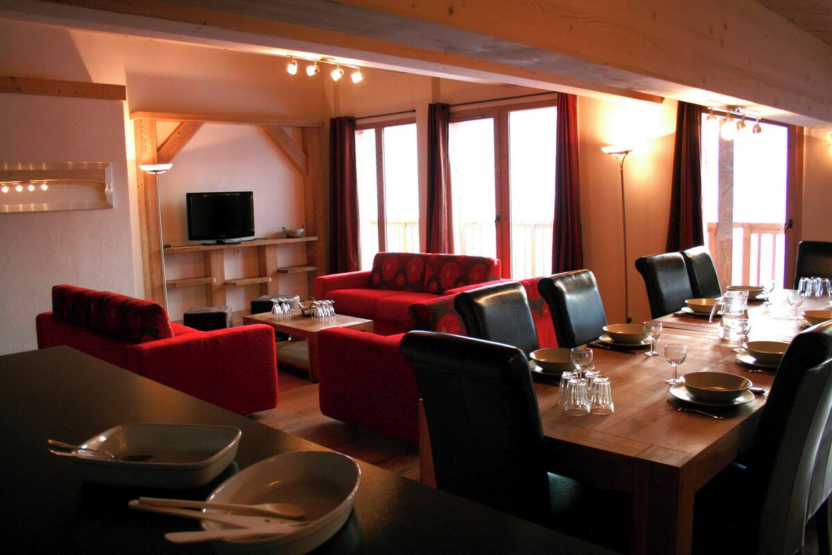 A dining area with a wooden table set for a meal, red upholstered sofas, and a television on a shelf.
