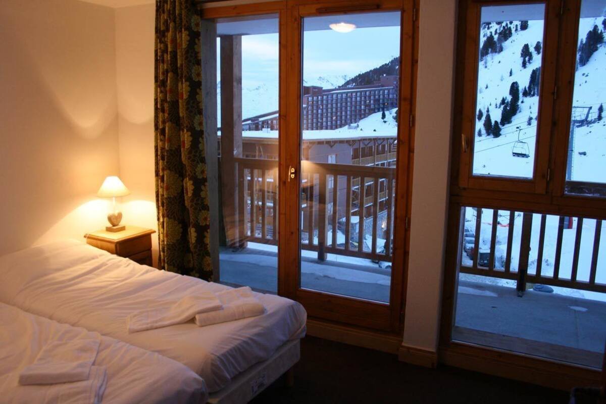 A hotel room with a bed, a nightstand with a lamp, and a balcony with a ski lift view.