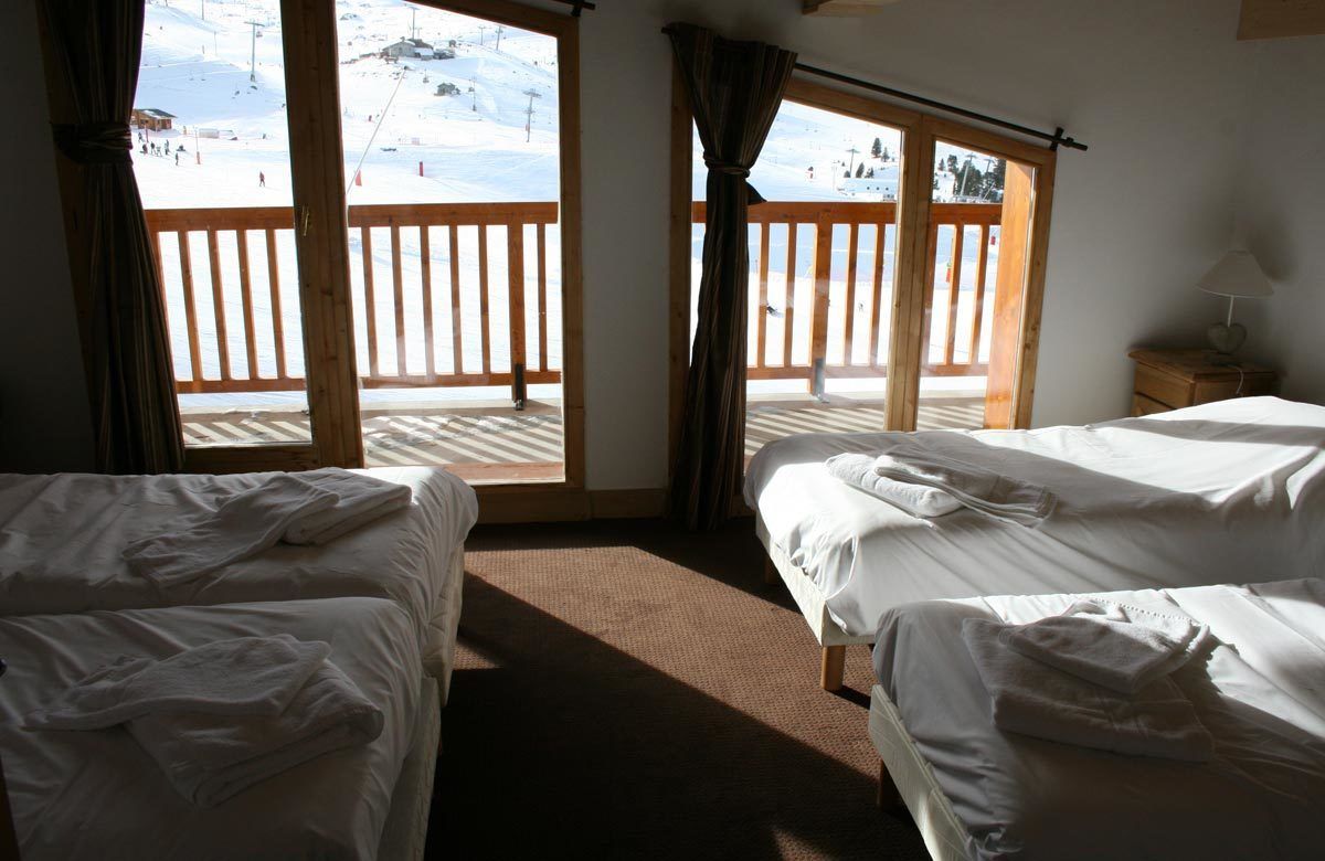 A hotel room with two beds, a wooden floor, a window with a view of a ski slope, and a nightstand with a lamp.