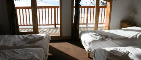 Habitación de hotel con dos camas, suelo de madera, ventana con vistas a una pista de esquí y mesita de noche con lámpara.