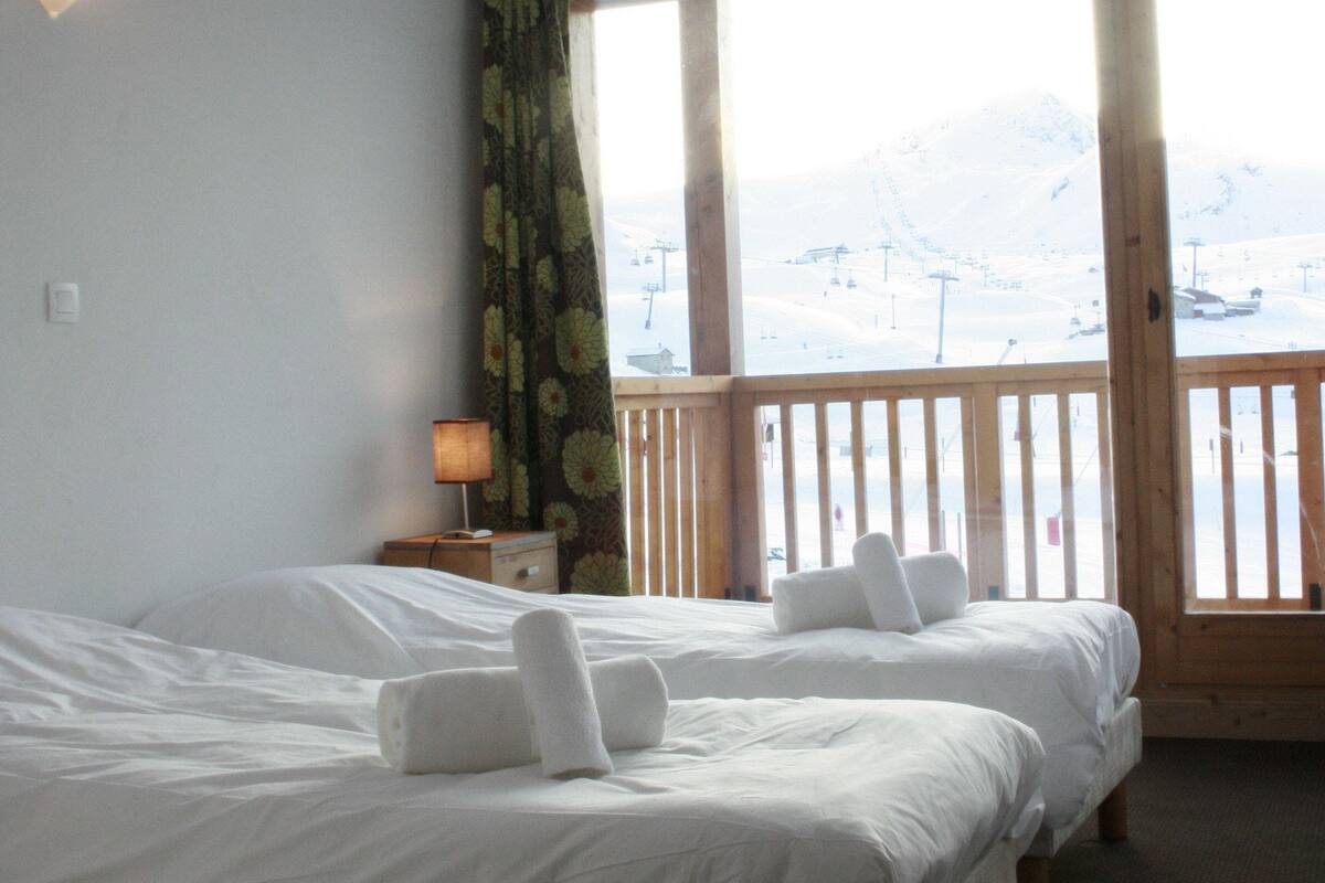A hotel room with two beds, a nightstand, a lamp, and a view of a snowy landscape through large windows.