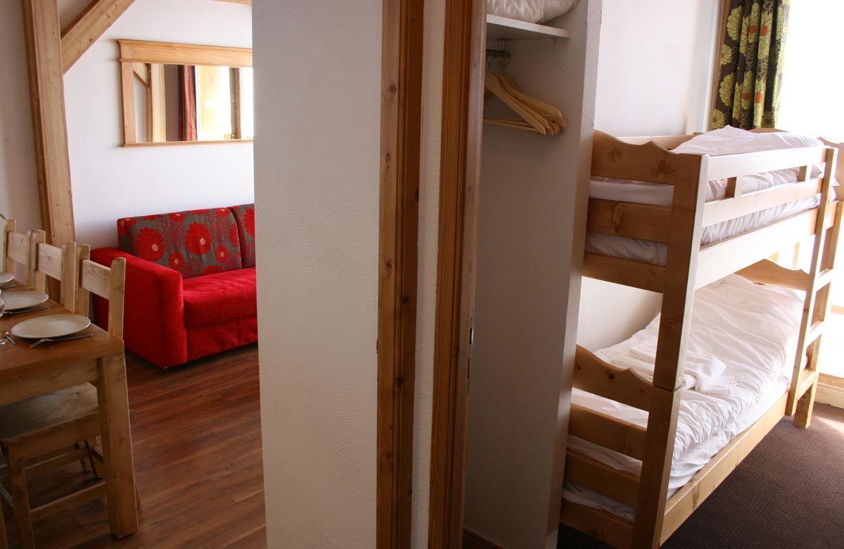 Una habitación con una cama litera, un sofá rojo, una mesa de madera con platos y un espejo en la pared.