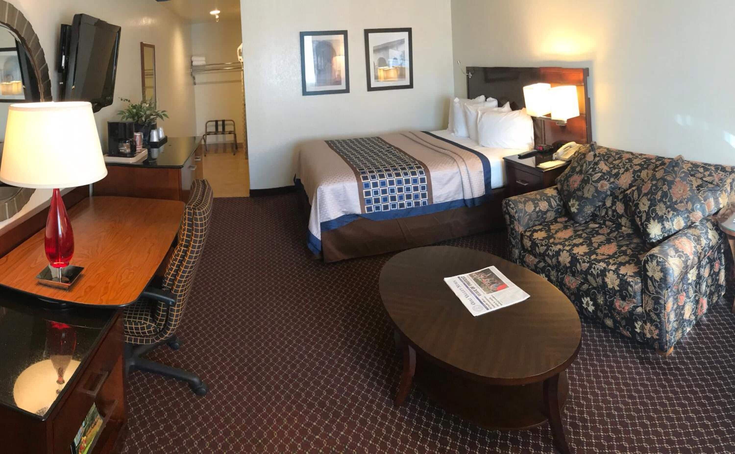 A hotel room with a bed, a sofa, a desk with a lamp, a chair, and a small table.