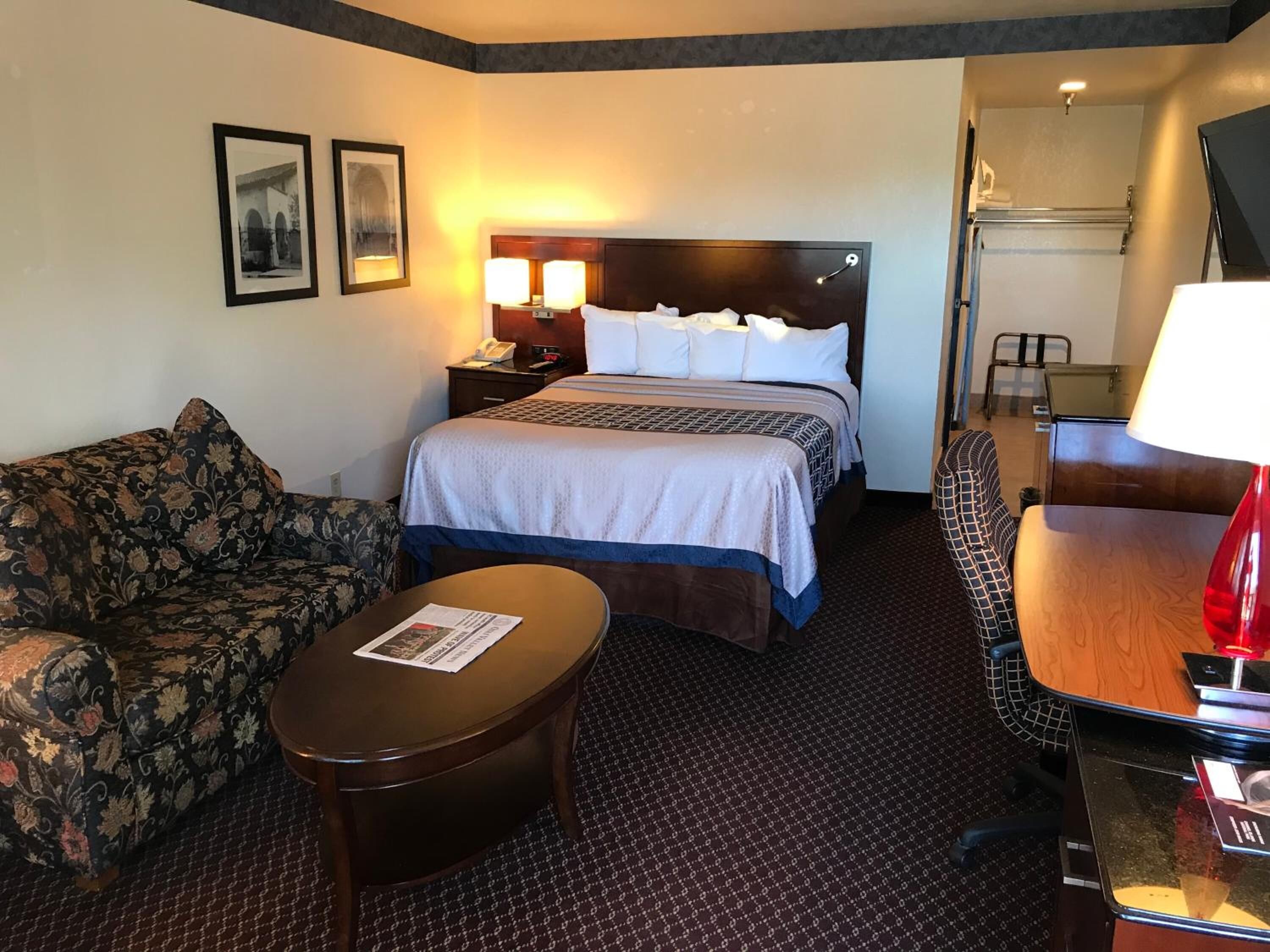 A hotel room with a bed, a sofa, a small table, and a desk with a lamp. There are two framed pictures on the wall.