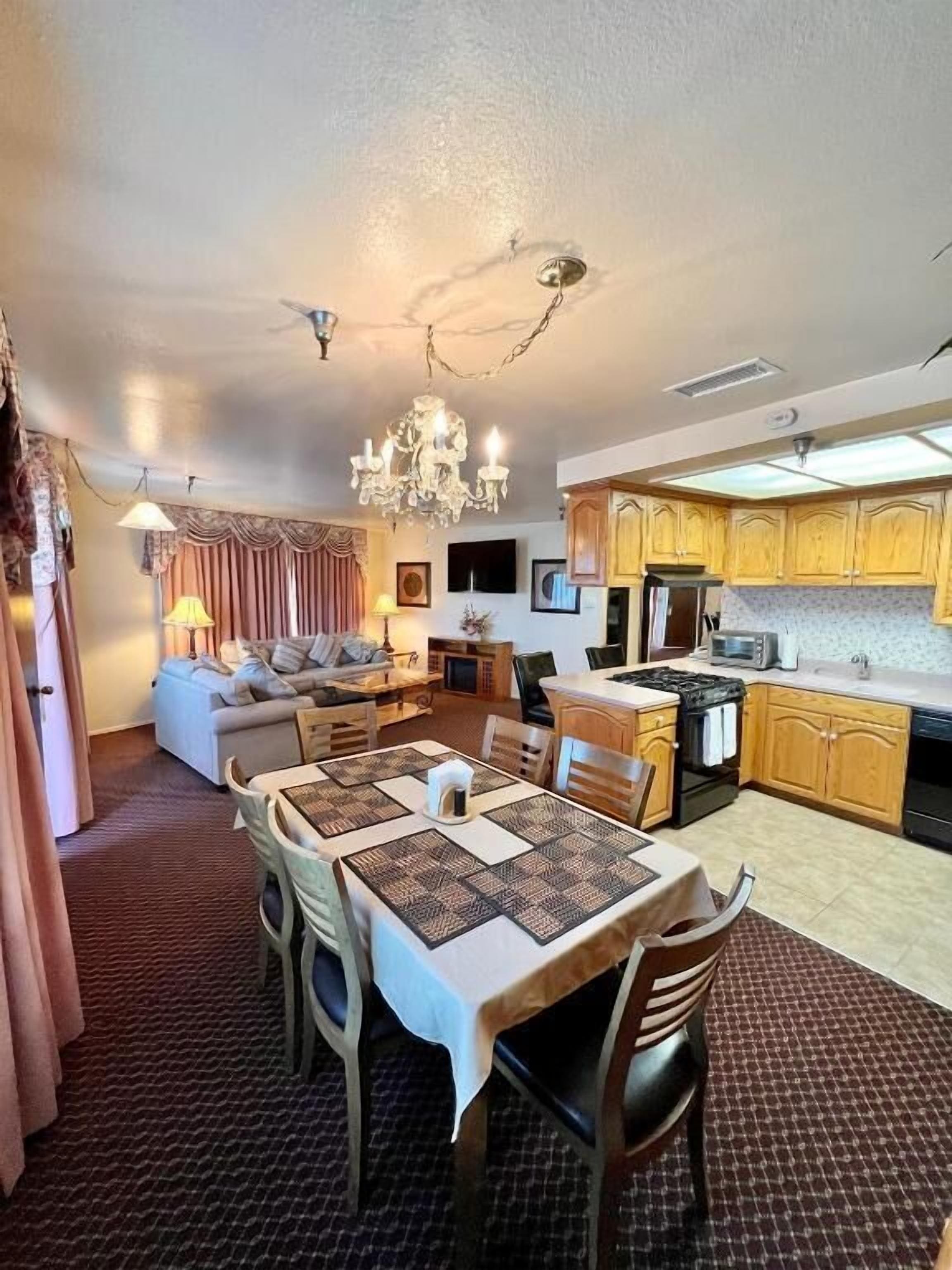 A dining area with a table set for four, a chandelier, and a kitchen in the background.
