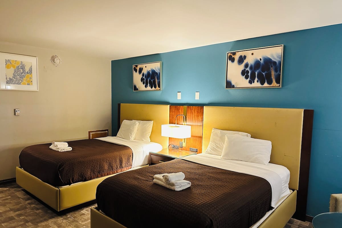 A hotel room with two beds, a nightstand, a lamp, and artwork on the walls.