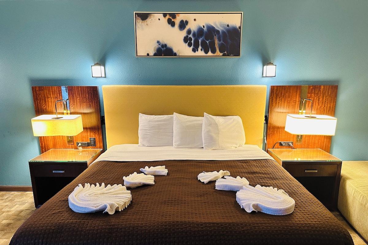 A hotel room with a large bed, two bedside lamps, a painting on the wall, and towels arranged on the bed.