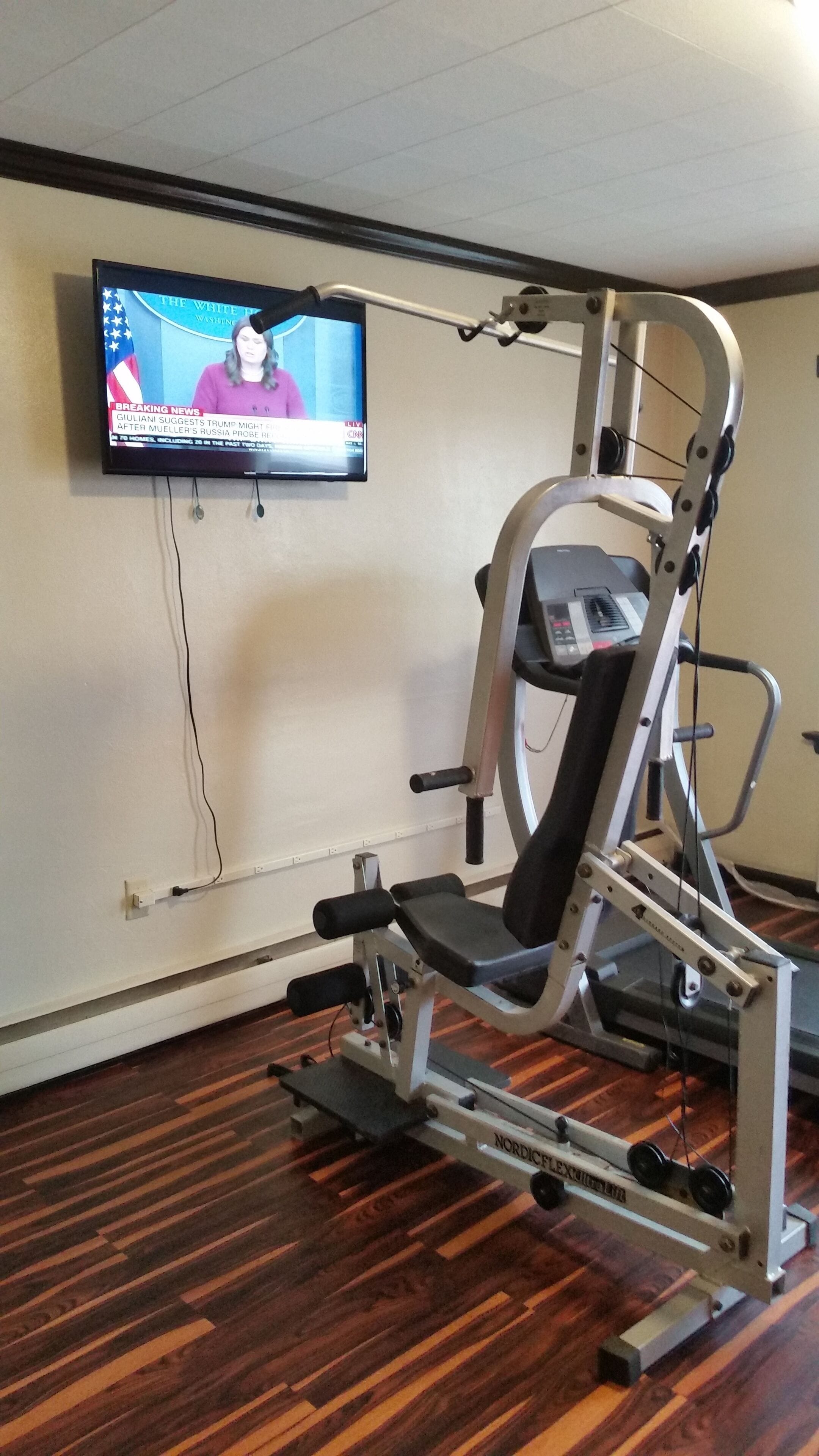 A gym with a weight machine and a television displaying news on the wall.