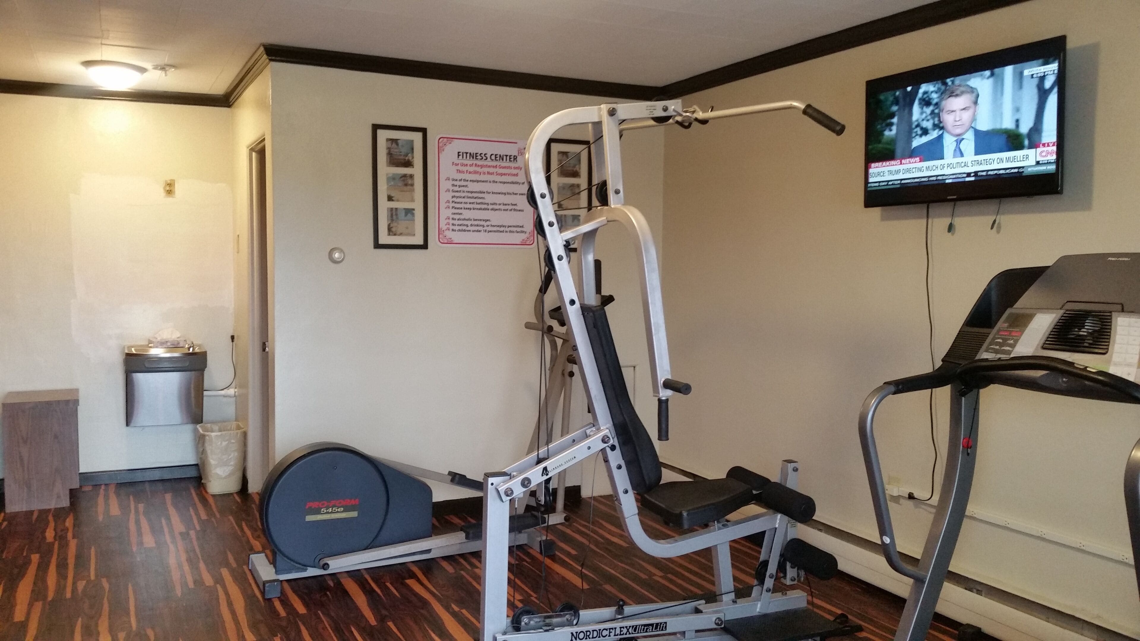 Une salle de sport équipée de machines de cardio, d’un téléviseur fixé au mur et d’un panneau indiquant les règles d’utilisation.
