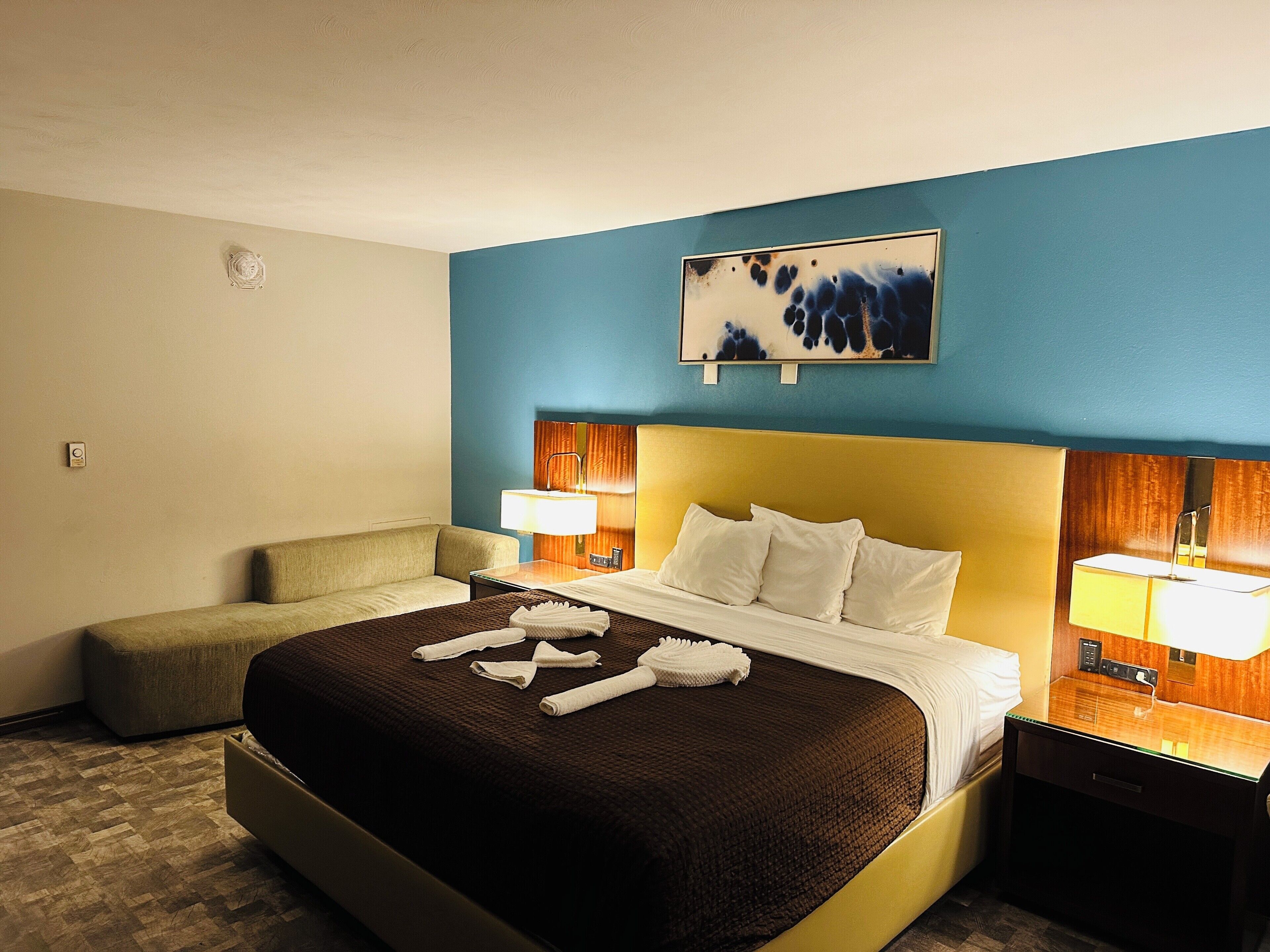A hotel room with a large bed, a bedside table with a lamp, a small sofa, and a painting on the wall.