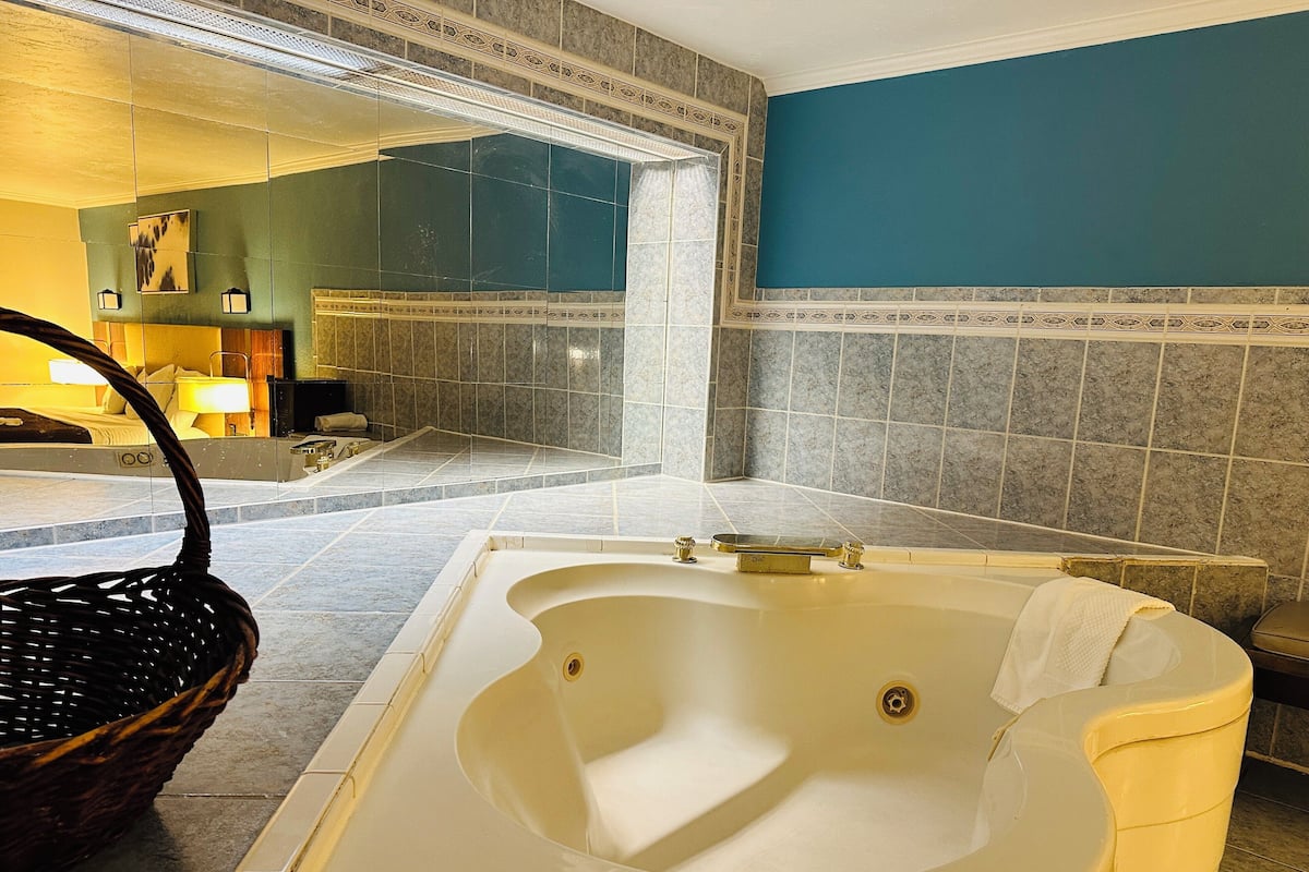 A bathroom with a large bathtub, a wicker basket, and a mirror reflecting the room.