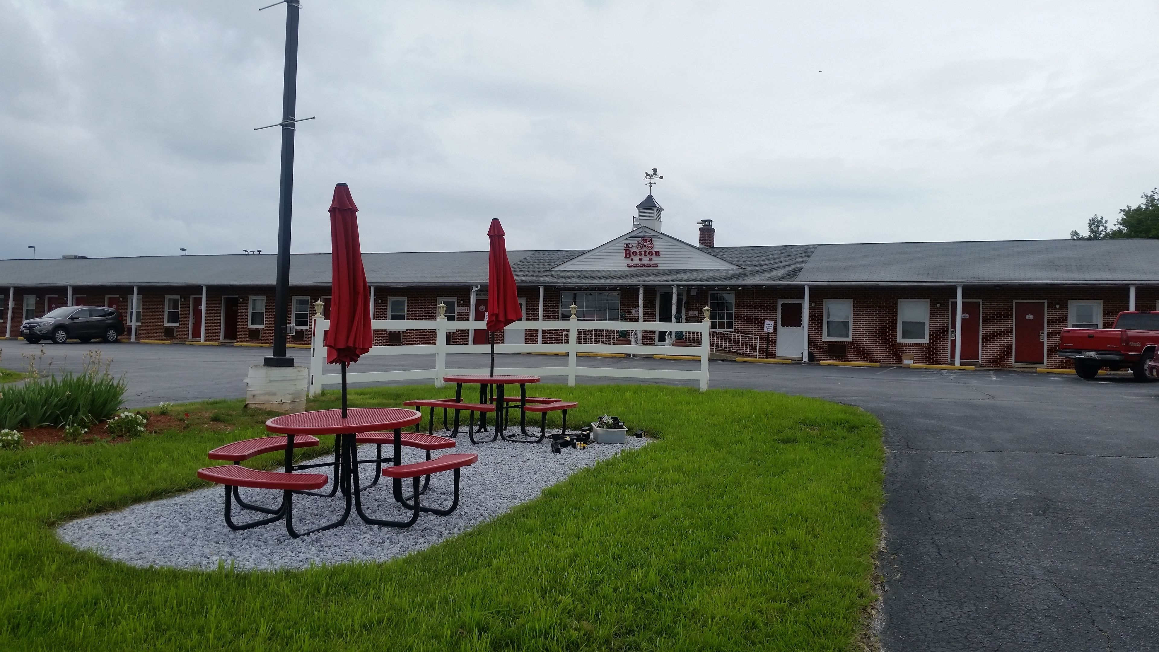 A motel with a red-brick exterior, a white fence, and a covered outdoor seating area with red tables and umbrellas.