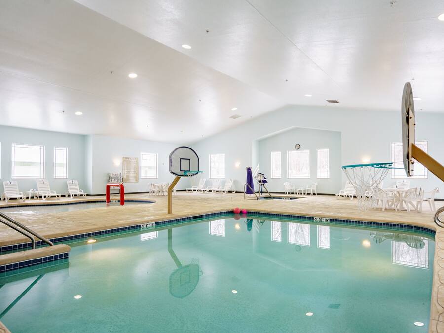 An indoor swimming pool with clear blue water, surrounded by white lounge chairs and a basketball hoop.