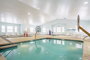An indoor swimming pool with clear blue water, surrounded by white lounge chairs and a basketball hoop.