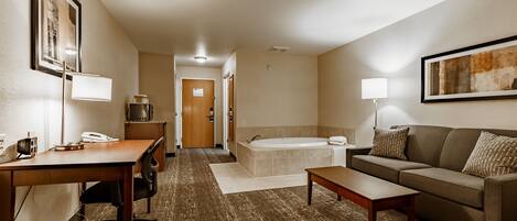 A hotel room with a wooden desk, a sofa, a coffee table, a bathtub, and a framed picture on the wall.