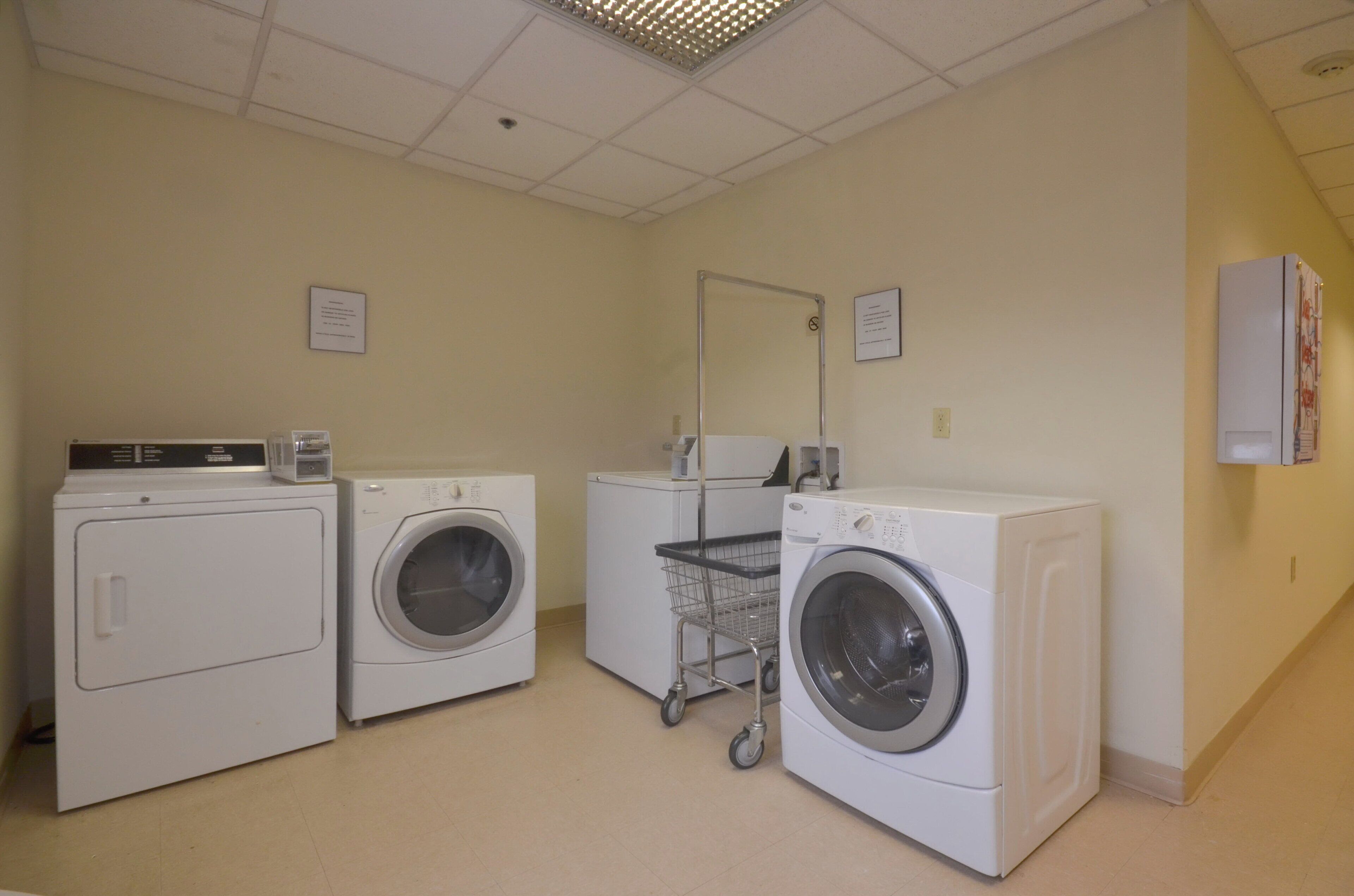 A laundry room with three washers and dryers, a cart, and two informational signs on the walls.