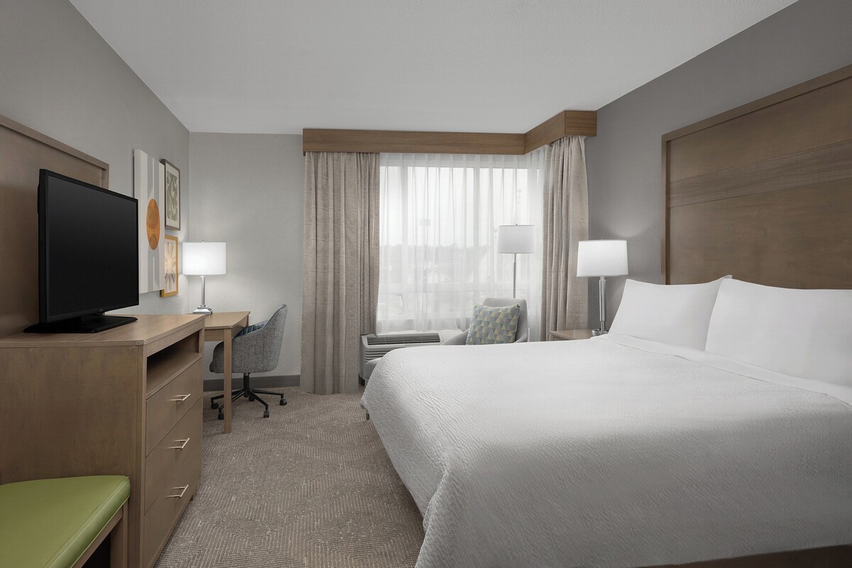 Een hotelkamer met een bed, een bureau, een televisie, een stoel en een raam met gordijnen.
