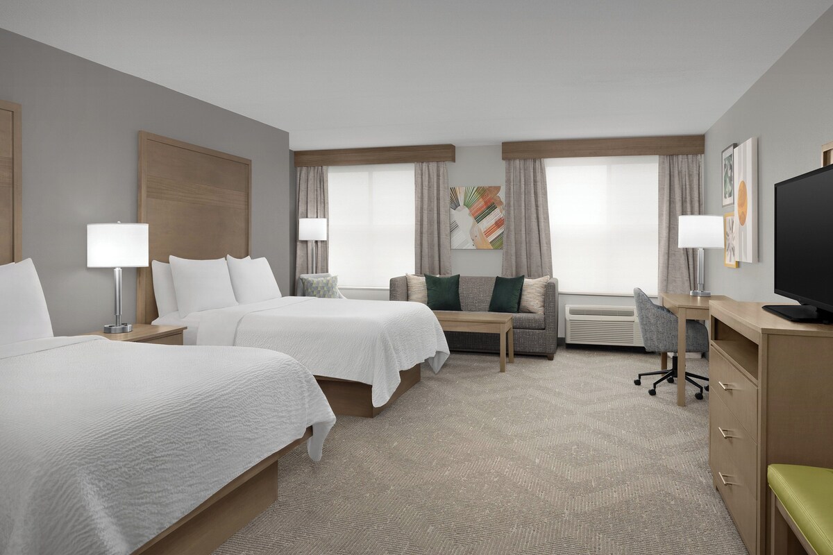 Een hotelkamer met twee bedden, een slaapbank, een bureau, een televisie en een groot raam met gordijnen.