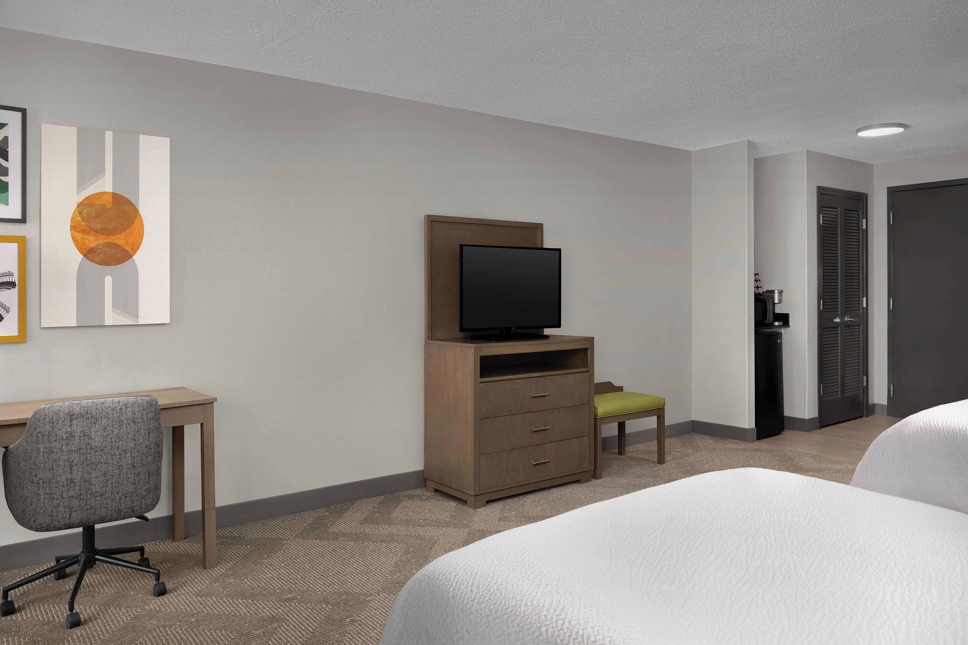 A hotel room with a bed, a desk with a chair, a television on a wooden cabinet, and a door to the right.