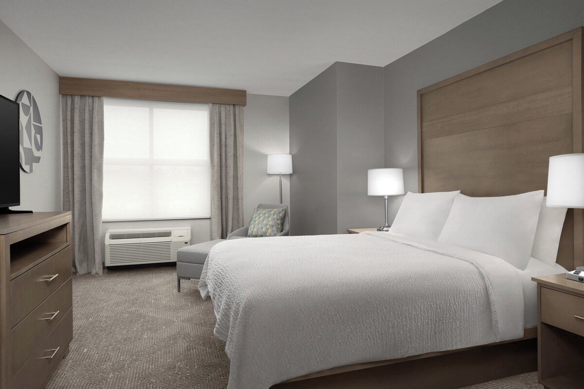 A hotel room with a bed, bedside tables, a desk, a chair, a TV, a window with curtains, and an air conditioning unit.