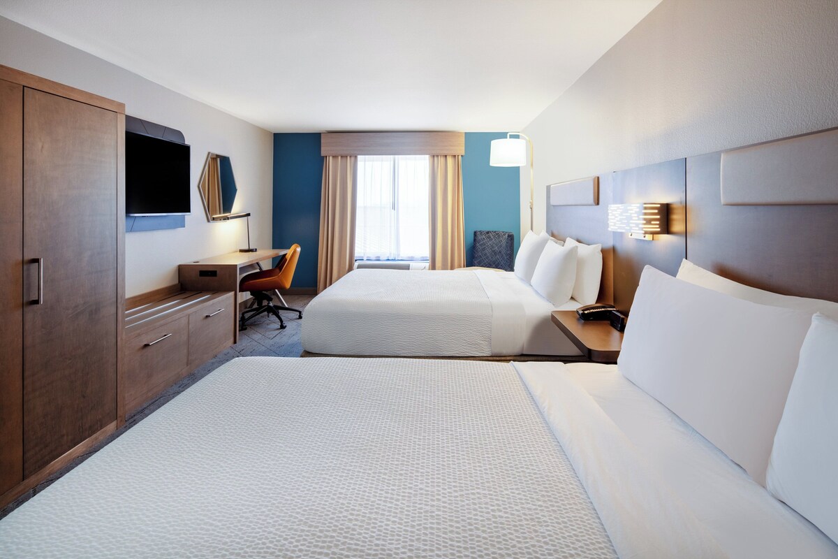 Ein Hotelzimmer mit zwei Betten, einem Schreibtisch mit Stuhl, einem Fernseher und einem Fenster mit Vorhängen.